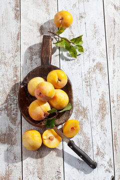 Sliced and whole yellow gages (Prunus domestica subsp. italica) and a wooden spoon on white wooden table, studio shot
