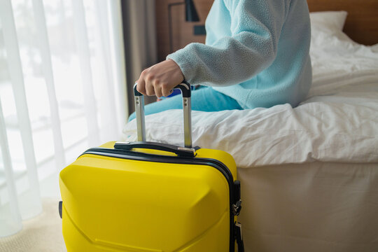 Side view of a male traveler holding luggage handle while resting on a couch or bed near window. Concept of travel arrival, hotel check-in, cozy tourism accommodation, and departure waiting.