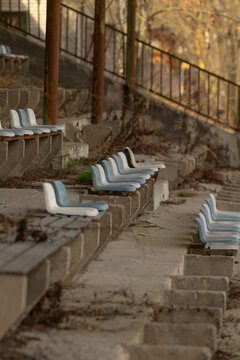 Rows of weathered white plastic seats fixed to cracked concrete terraces in an old outdoor sports stand. Rusted metal railings and vertical support posts run