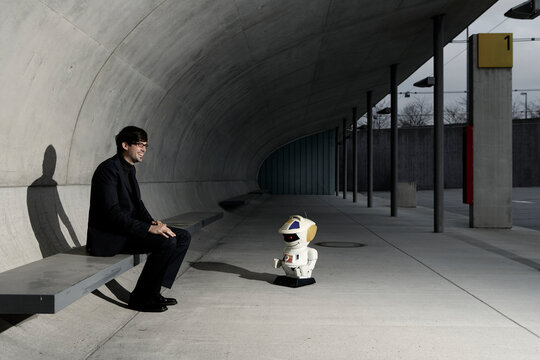 Laughing businessman sitting with a toy robot on a modern platform