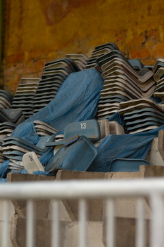 A pile of old blue plastic stadium seats stacked and disassembled in a storage area. Many numbered seat backs and bases are visible, showing scuffs, scratches