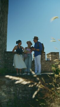 A group of tourists explores a historic stone wall, checking a map and mobile phone for their route. Three friends enjoy a sunny day of vacation travel and sightseeing.