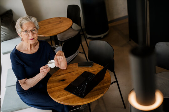 Smiling senior woman enjoying coffee and laptop at caf� table indoors
