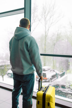 Cropped view of a male traveler holding luggage handle near large window with winter background. Concept of travel arrival, departure, hotel check-in, and winter holiday trip.