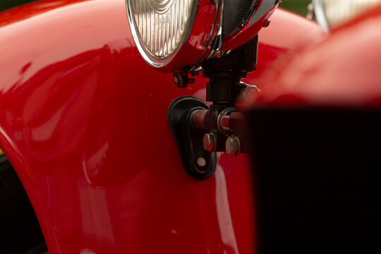 Tight detail of a red vintage car showing a chrome headlight assembly mounted above a glossy curved fender. Visible are metal brackets, a small mounting hinge