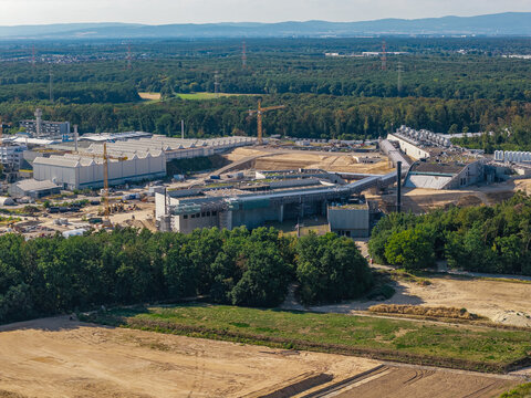 Aerial view of the Facility for Antiproton and Ion Research construction site with cranes, industrial buildings, and surrounding forest in Darmstadt, Hessen, Germany.