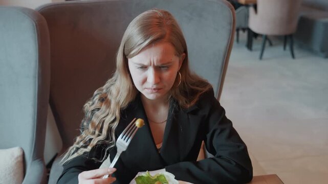 Young woman grimacing at bitter salad, modern cafe setting, black blazer, long hair, forkful greens rejected, facial frown, body language shows displeasure, subtle cinematic tones