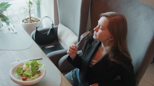White woman tasting salad in cafe, business executive pauses between bites seated in plush chair by window with handbag, fresh lettuce tomato cucumber in ceramic bowl, natural light, elegant black
