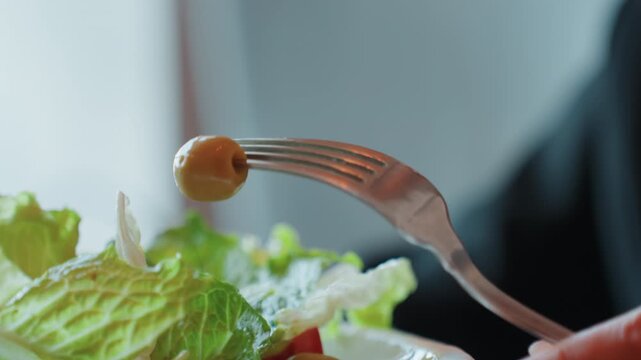 Closeup fork holding olive over salad, crisp romaine leaves glistening with dressing, hand guiding fork toward bowl, soft natural light, casual indoor dining, focus on texture and freshness
