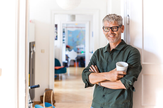 Portrait of smiling mature man with coffee cup leaning against door case at home