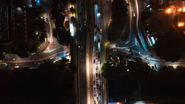 Aerial view of a busy highway intersection at night with illuminated traffic lanes, street lights, and surrounding foliage in Jakarta, Indonesia.