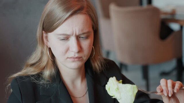 Woman tasting spoiled lettuce in cafe, office manager in blazer inspects wilted leaf on fork with growing grimace, covers mouth in queasy reaction, subtle restaurant interior, candid closeup capturing