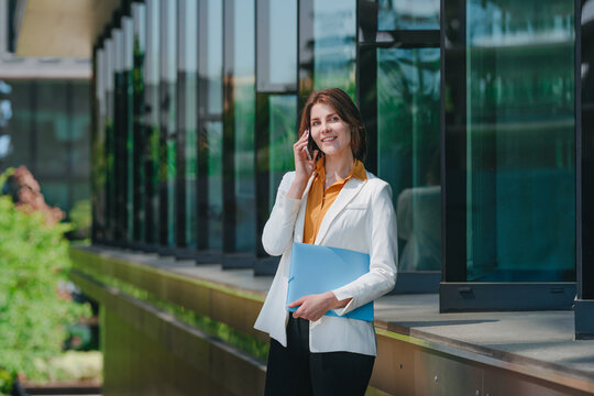 Smiling businesswoman talks on smartphone outdoors holding a folder