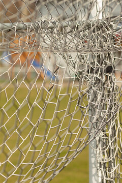 Detailed close-up of a damaged metal chain-link fence and bent support post at an outdoor field. The image shows twisted steel wire, broken ties