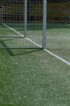 Close-up view of a metal soccer goalpost and net anchored on green artificial turf near the white sideline. Sunlight casts crisp shadows of the net and frame