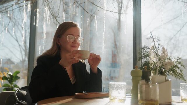 Caucasian woman smiling with coffee cup bright sunlight casting soft flare upbeat expression modern bistro interior pastry on plate glass of water blazer and glasses confident morning pause social