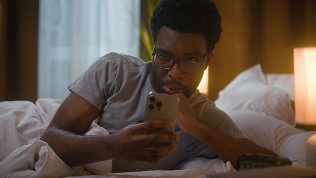 African American man resting lying on bed using mobile phone smartphone screen browsing internet watching online video content satisfied relaxed ethnic male user client blanket at home bedroom evening
