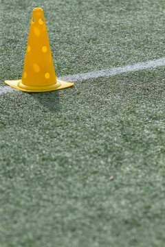 A bright yellow plastic training cone with circular holes sits upright on green artificial turf next to a painted white field line. The shallow depth of field