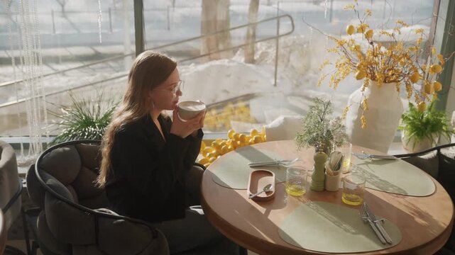 Sunlit cafe table white woman sipping coffee and tasting pastry, floral vase centerpiece, ceramic cup and spoon, placemat and journal, soft window light, warm golden tones, relaxed contemplative