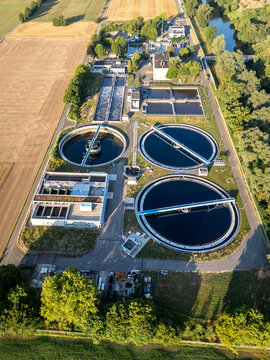 Aerial view of a waste water treatment plant with circular sedimentation tanks and rectangular aeration basins surrounded by agricultural fields and lush trees in Germany.