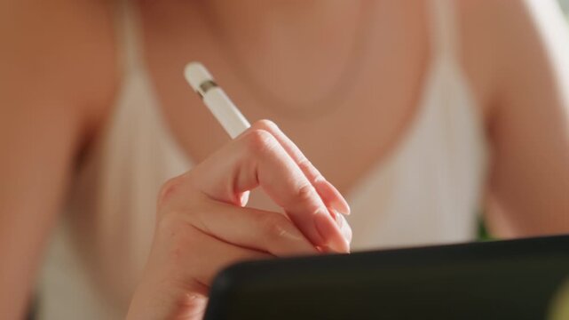 Closeup hands drawing on tablet with stylus creating fine strokes and composition, blurred torso in white top and necklace visible, warm natural light, focused creative mood and studio detail