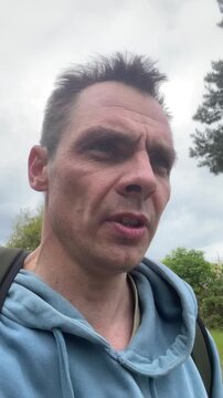 Close-up vertical portrait of a middle-aged man outdoors adjusting the right side of his light blue hoodie while looking slightly downward at the camera. He
