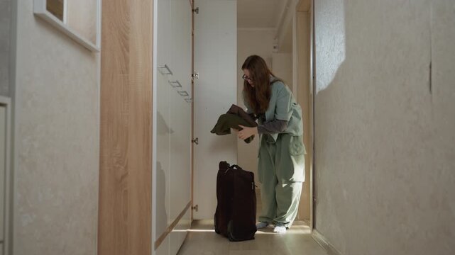 Caucasian woman secures front door with suitcase purposeful commuter unlocks latch hangs coat seals suitcase checks pocket rolls baggage toward threshold sunlit foyer confident relief forward motion