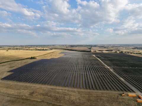 Aerial view of a large solar farm with rows of photovoltaic panels set in a rural landscape with dry fields and a blue sky with clouds at Irchester, England, United Kingdom.