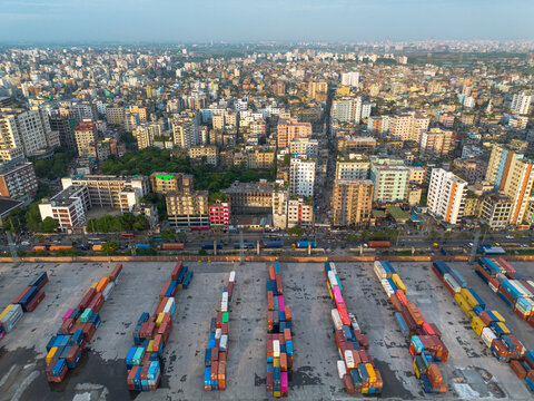 Aerial view of a busy container terminal with rows of colorful shipping containers in the foreground, set against the dense urban skyline of Dhaka, Dhaka Division, Bangladesh.