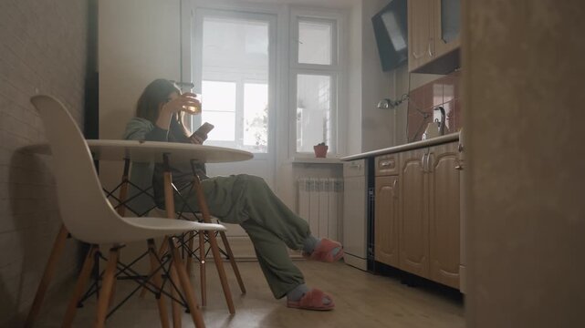 Young woman reclining kitchen chair phone, slouched posture scrolling messages, late afternoon light through window, slippers and pajamas, mug on table, freelance fatigue and daydreaming,