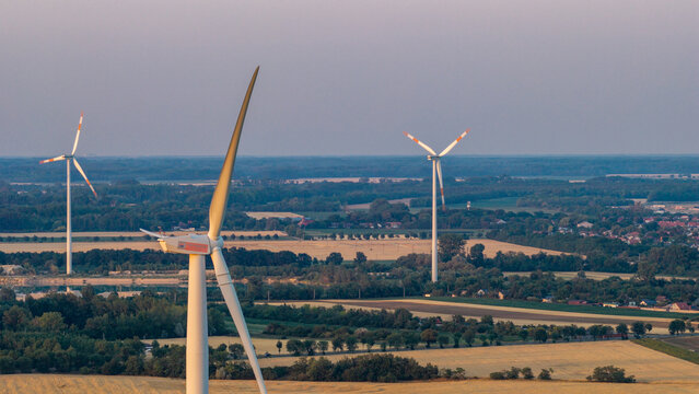 Aerial view of wind turbines in a rural landscape with golden fields, trees, and a small town in the distance under a soft sky in Level, Hungary.
