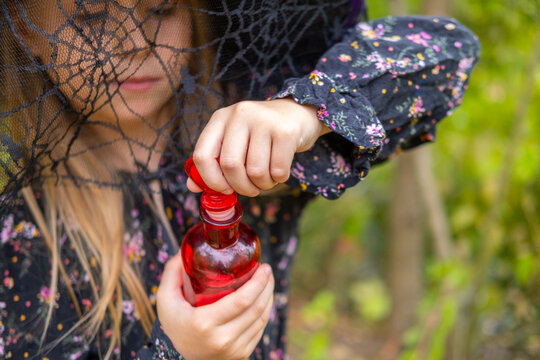 girl in black witch hat holding red glass potion bottle with heart stopper, little sorceress preparing love spell in magic forest, first love, cute childhood fantasy story, pagan witchcraft ritual