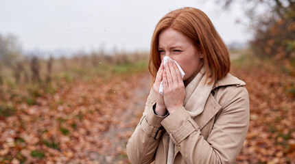 Obraz premium Young woman with red hair blowing her nose into a white tissue, surrounded by fallen autumn leaves