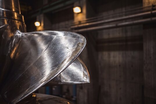 A detailed close-up of the massive, shiny stainless steel runner blades of a hydroelectric turbine inside a power station powerhouse.