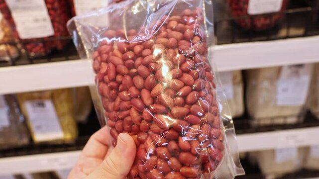 Red peanuts in transparent plastic package held in hand at grocery store. Close up of raw organic legume kernels in clear bag being selected by shopper from supermarket shelf display.