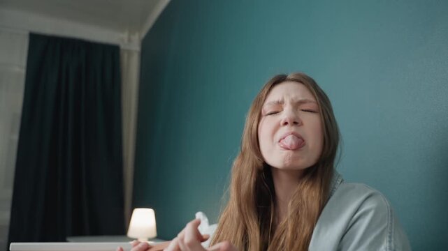 Young woman in bedroom making playful faces and tongue out, soft lamp glow and teal wall, candid laughter and quirky expressions, hands animated while interacting with unseen object, cozy intimate