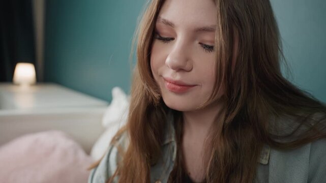 Caucasian girl sipping drink browsing tablet in cozy bedroom, soft lamp glow and amber beverage, thoughtful student winding down after study, long hair falling over shoulders, pajama top and gentle