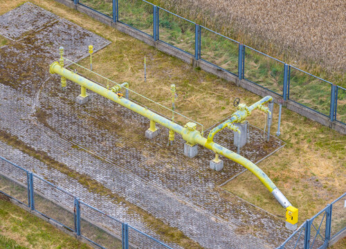 Aerial view of a yellow gas pipeline infrastructure situated within a fenced enclosure, surrounded by grassy terrain and agricultural fields in Slupsk, Pomeranian Voivodeship, Poland.