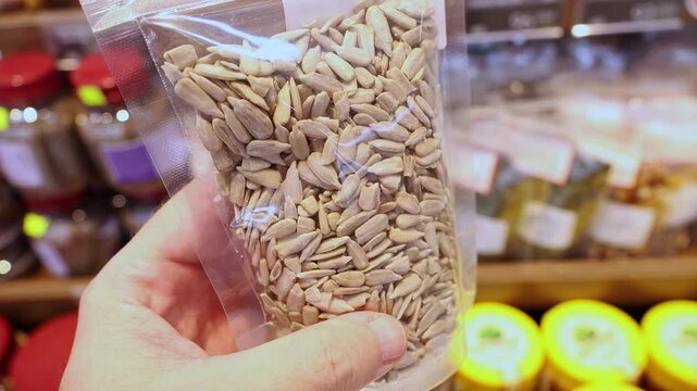 Sunflower seeds in transparent plastic package held in hand at grocery store. Close up of raw organic kernels in clear bag being selected by shopper from supermarket shelf display.