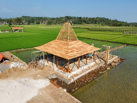 Aerial view of a traditional Javanese Joglo structure under construction, featuring a wooden roof frame set amidst lush green rice fields in Kecamatan Tingkir, Jawa Tengah, Indonesia.