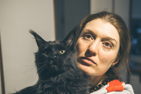 A young Caucasian woman with long brown hair holds a large black cat close to her face. The background is softly blurred, creating a cozy atmosphere.