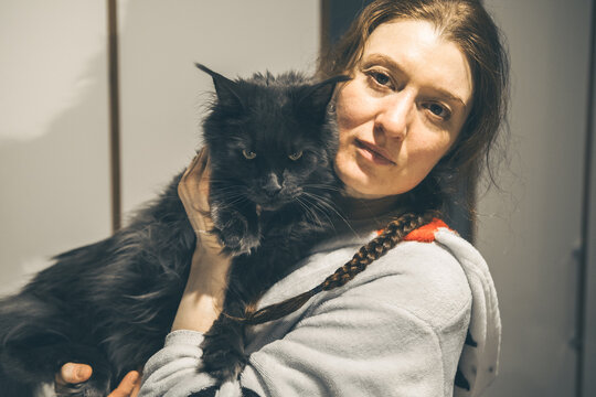 A young Caucasian woman with long brown hair holds a large black cat in her arms. The setting is indoors with neutral-colored walls.