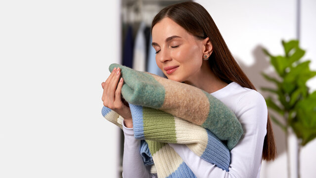 Young woman smelling clean wool sweaters, wardrobe and green houseplant on light gray home interior background. Concept fresh laundry, clothing knitwear care and seasonal wardrobe routine. Copy space