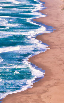 Turquoise ocean waves create white foam as they wash ashore onto a wide sandy beach next to dramatic eroded cliffs under a clear sky. San Mateo County, California, United States.