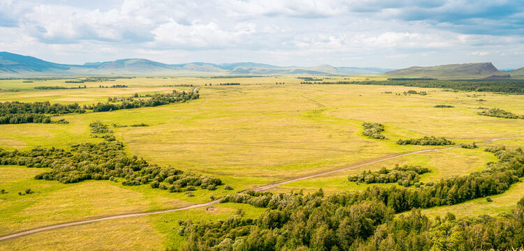 Aerial photography from drone, steppe valley with yellow-green meadows, dirt road, groves of bushes, gently rolling mountains on horizon, cloudy sky