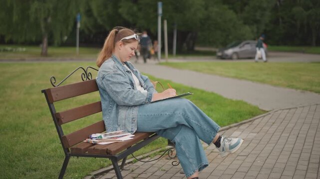 Young woman sitting on bench sketching journal, denim jacket and headband, watercolor set and pen beside, relaxed posture, treelined park and paved pathway, distant pedestrians and parked car, soft