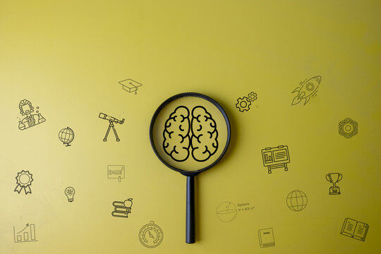 STEM education and creative thinking concept. Magnifying glass focusing on a digital brain graphic surrounded by science and learning icons on a yellow background with copy space.