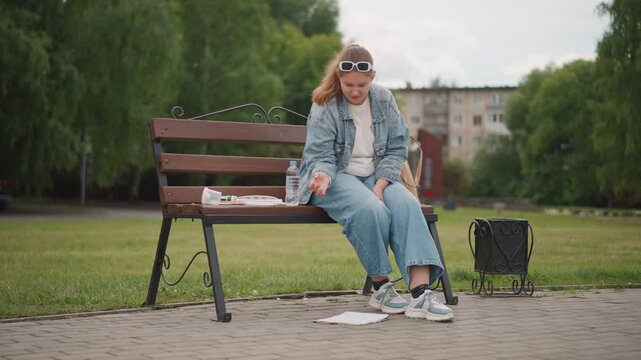 Young white woman on bench sorting papers and sketchbook, denim jacket and sneakers, sunglasses on head, backpack at side, wind scattering notes onto pavement, pensive expression showing study stress
