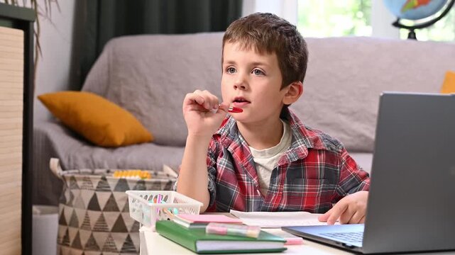 Boy practices writing homework while watching educational content on laptop, creating balanced learning routine that combines technology with traditional handwriting skills