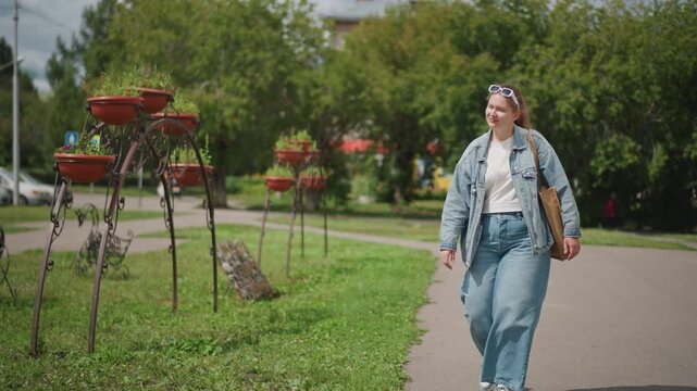 Young woman walking with shopping bag across park path, denim outfit, planters and parked cars in background, casual shopper stroll, candid daylight scene, bench rest ahead, peaceful city greenery